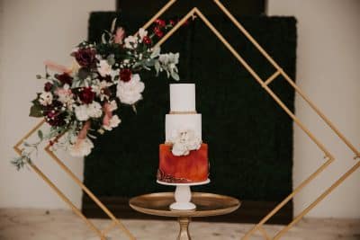 three tier cake on a cake stand, square metal design behind, flowers added to the side, bottom layer of the cake is red, other layers are white, Orlando, FL