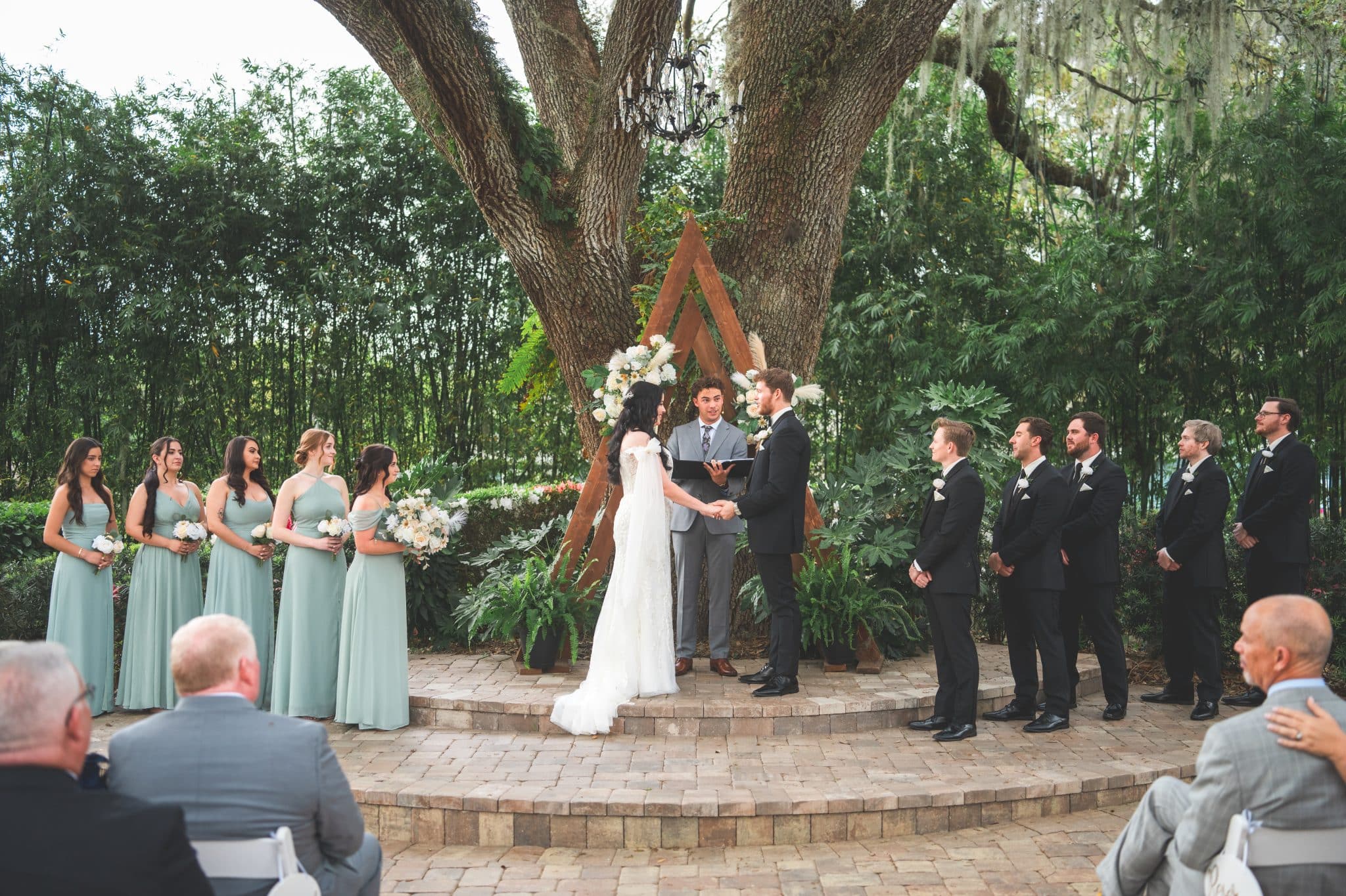 bride and groom during ceremony with bridal party in front of a forest backgroud with wooded arch and white floral arrangements