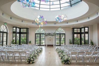 wedding ceremony set up in the conservatory, open atrium, colorful decor hanging from the ceiling, rows of chairs with white floral bunches at the foot of each row in the aisle, Orlando, FL