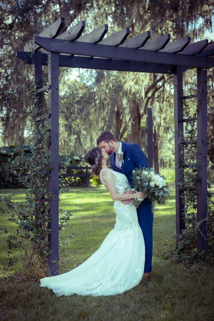 bride and groom standing under a pergola, groom kissing and dipping his bride, Dream Day Digital, Orlando, FL