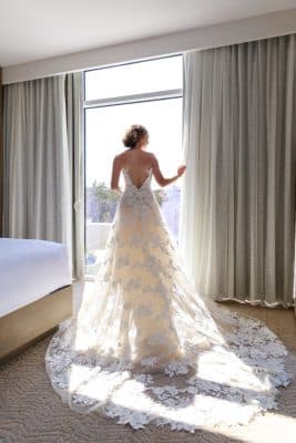bride in her wedding dress, sheer lace train spread behind her, standing in a hotel room facing the window, Orlando, FL