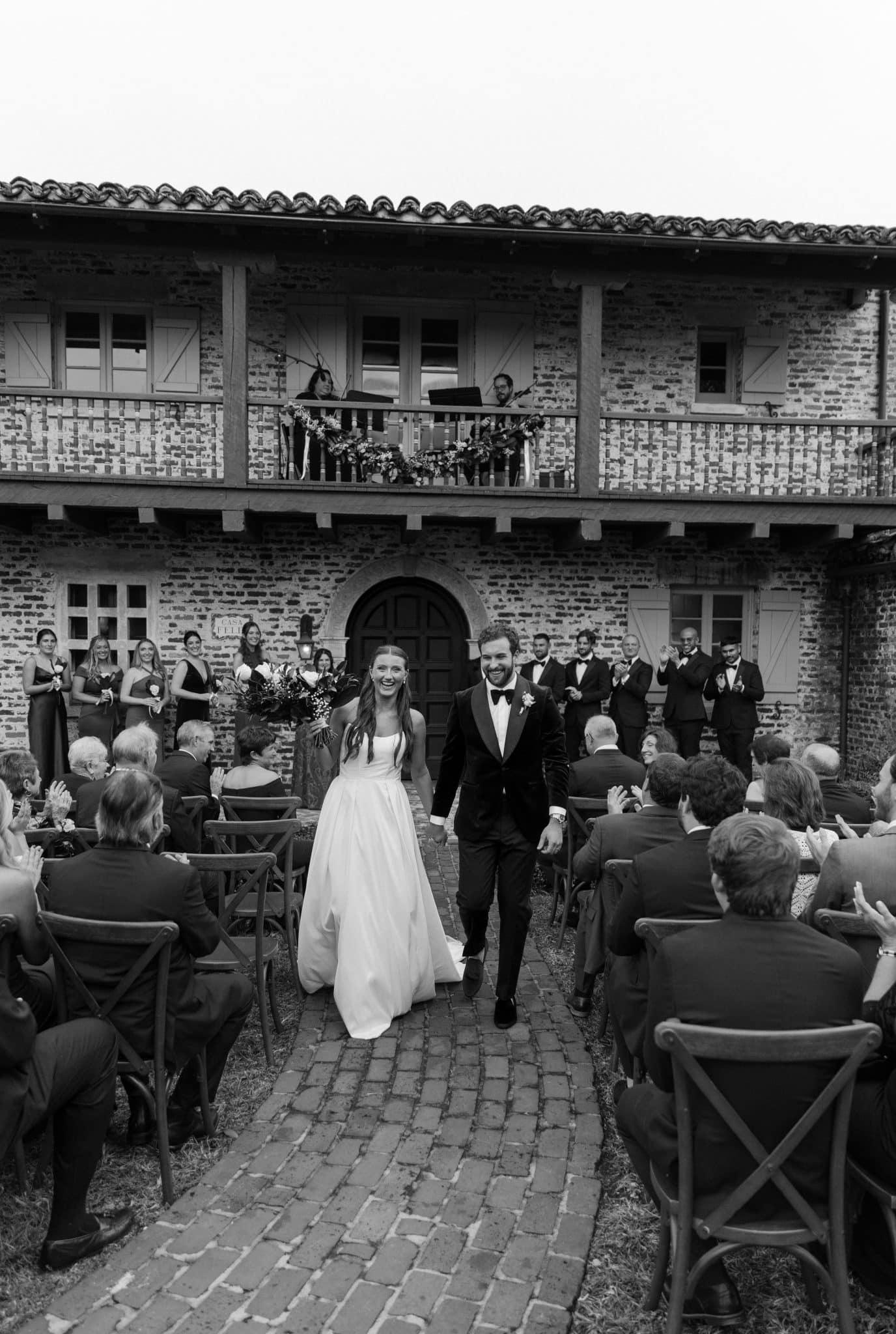 black and white image of a couple walking down the aisle with a live musician playing on the second story behind them 