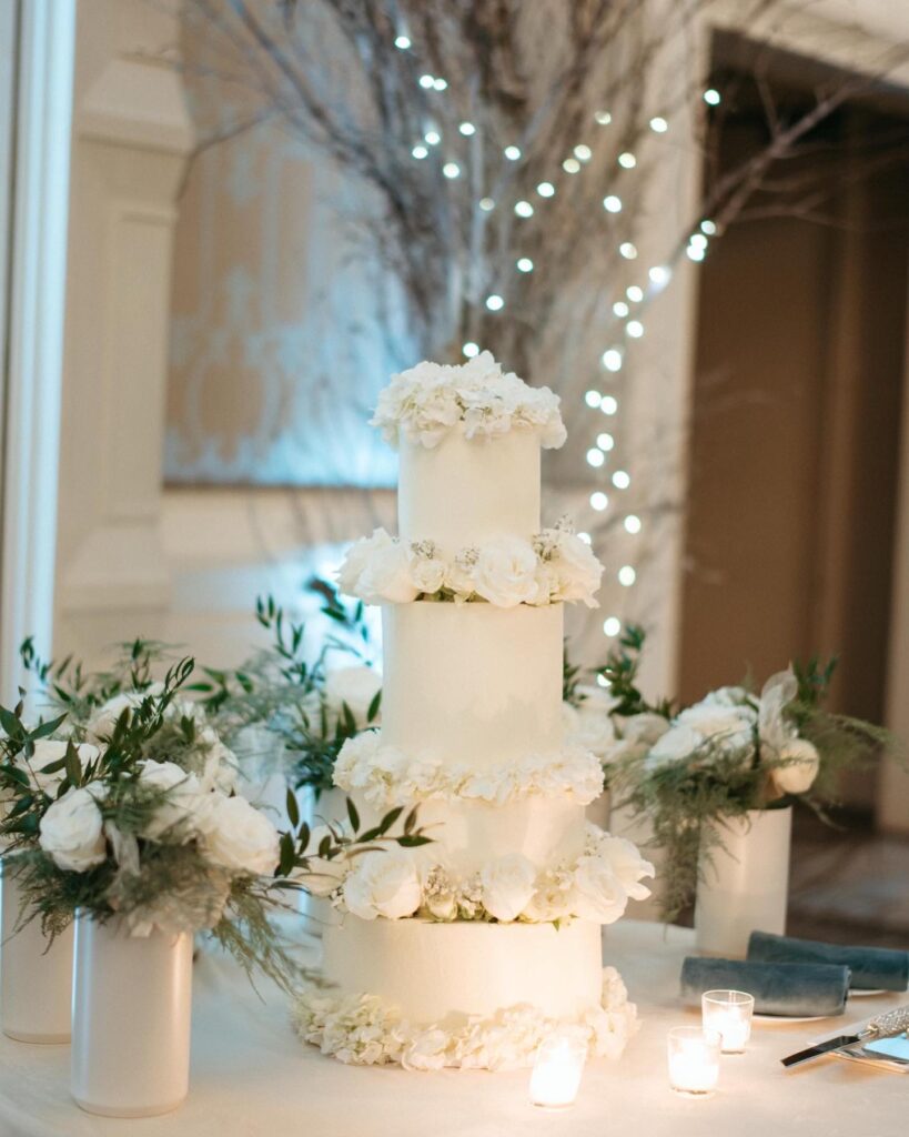 four tiered white cake adorned with white flowers, white vases on the left and right of the cake, filled with white flowers and greenery, Iced by Taylor, Orlando, FL