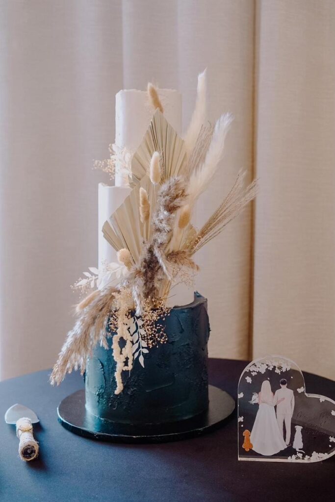 tiered wedding cake with top layers being white and bottom being teal, cake server laying on the table, decorated with dried flowers and an acrylic hear design on the table, painted with the wedding couple, Iced by Taylor, Orlando, FL