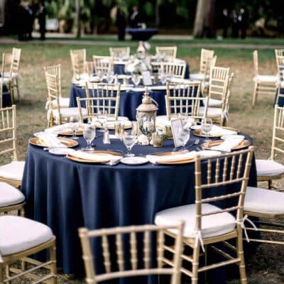 large round table with a navy blue tablecloth, gold chargers, white napkins, white cushions on gold chairs, outdoors, Orlando, FL