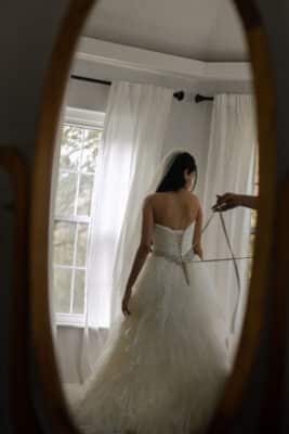 bride having her dress secured in the back, viewed through the oval mirror in the room, Orlando, FL