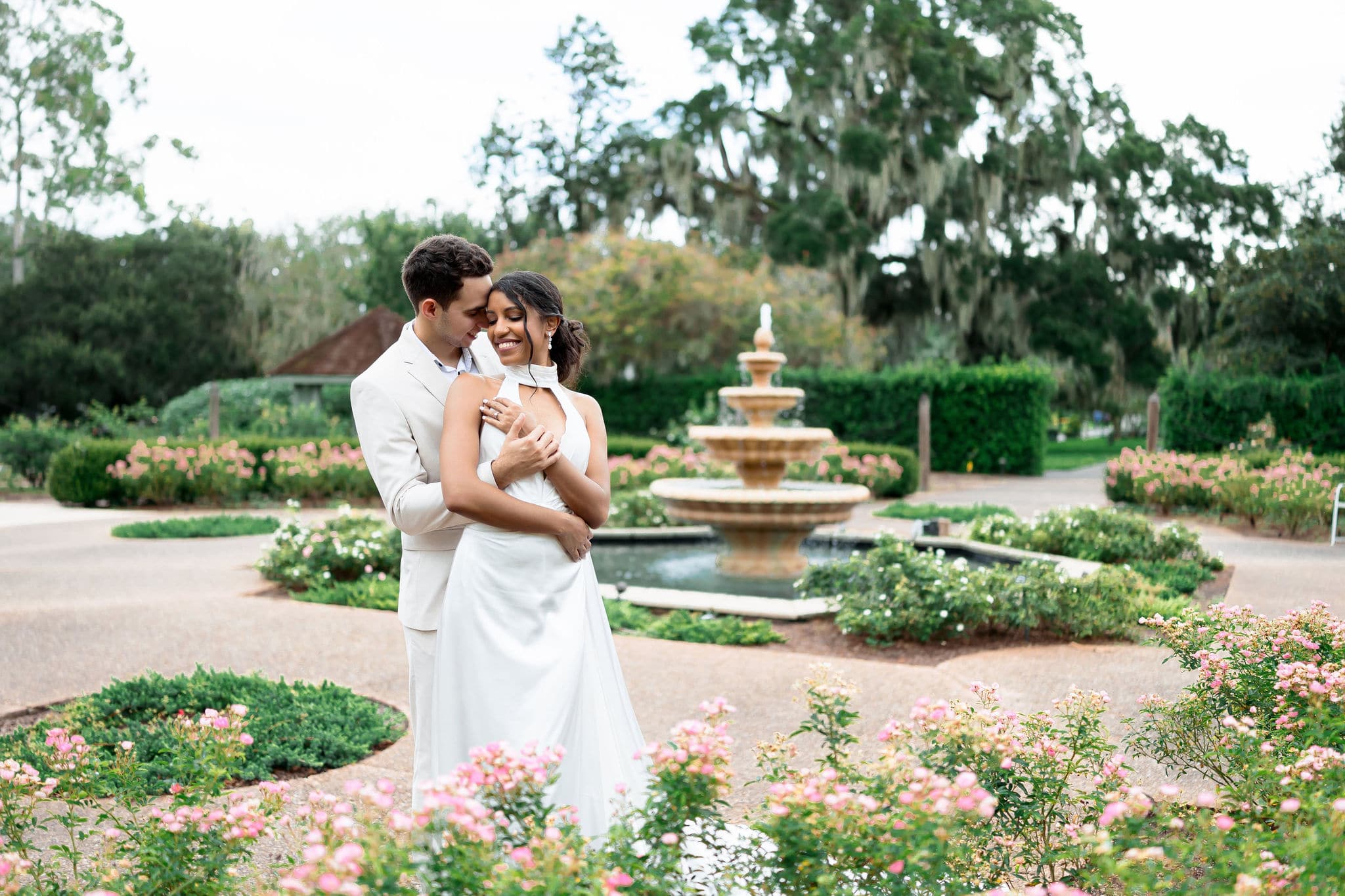 bride and groom both wearing white in front of a fountain with gorgeous greenery and pink flowers surrounding them