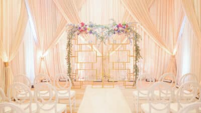 Elegant indoor wedding ceremony setup at Lake Nona Wave Hotel in Orlando Florida featuring a gold geometric arch adorned with colorful floral arrangements and greenery, white ghost chairs, and soft blush pink draping throughout the ballroom