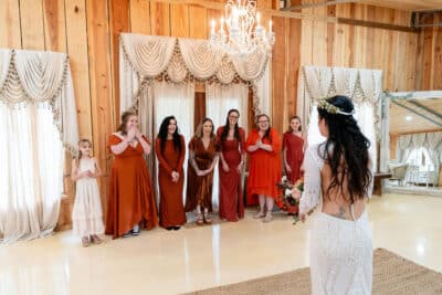 bride dressed in her gown, facing her wedding attendants in red dresses, flower girl in a white dress, Rustic Grace Barn, Orlando, FL