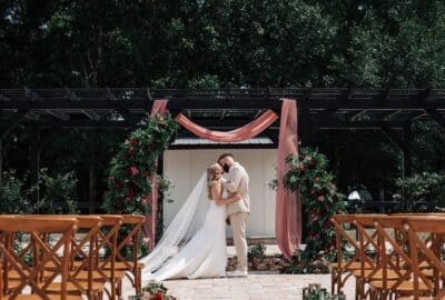 bride and groom kissing at the altar, wedding ceremony, wooden chairs, perfotal draped with pink sheer fabric and greenery, Orlando, FL
