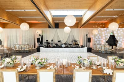 Elegant wedding reception setup at Bonnet Springs Park in Lakeland Florida featuring a white dance floor, round paper lantern chandeliers, draped fabric stage backdrop with live band setup, wooden farm tables with white floral centerpieces, and warm wood beam ceiling in the modern ballroom event center