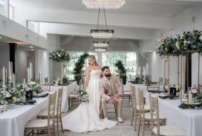 Bride and groom pose together in the elegant white reception hall at Villa Serafina wedding venue in Central Florida, surrounded by gold chiavari chairs, white linen-draped tables, tall floral centerpieces with greenery, and crystal chandeliers