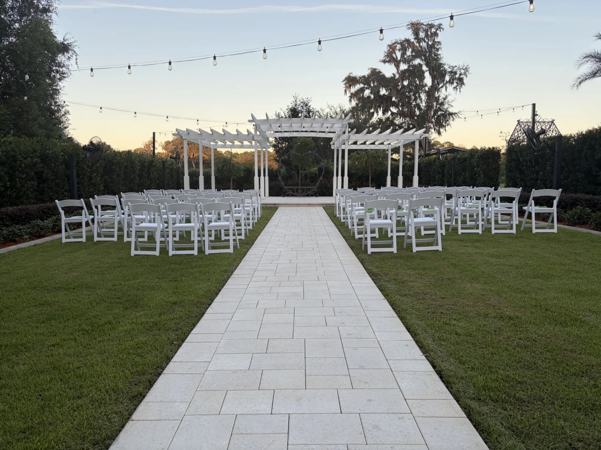 Outdoor wedding ceremony setup at Ruby Oaks Events in Central Florida featuring a white pergola altar, white folding chairs lining a stone aisle, and string lights overhead at sunset