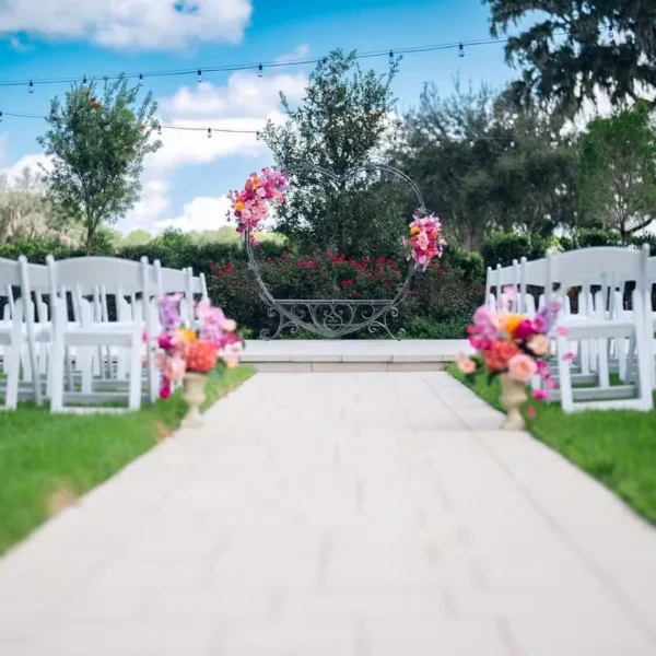 Outdoor garden wedding ceremony setup at Ruby Oaks Events in Central Florida featuring a circular floral arch with pink and orange blooms, white folding chairs lining a wood plank aisle, and colorful floral arrangements on pedestals under a blue sky