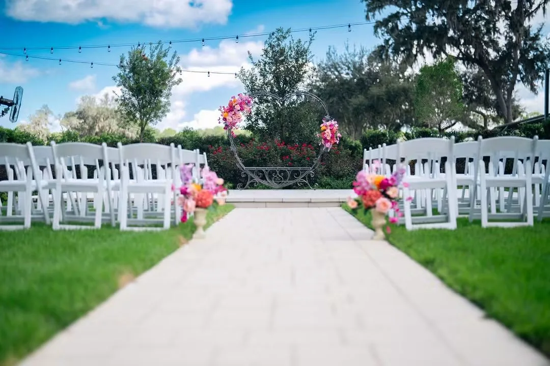 Outdoor garden wedding ceremony setup at Ruby Oaks Events in Central Florida featuring a circular floral arch with pink and orange blooms, white folding chairs lining a wood plank aisle, and colorful floral arrangements on pedestals under a blue sky