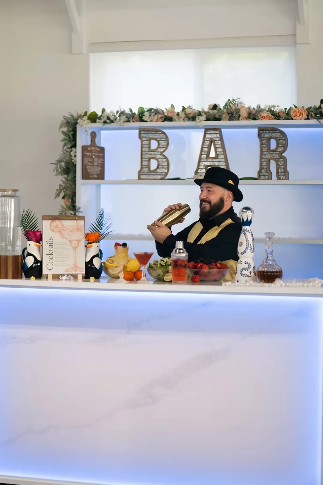 Smiling bearded bartender in a black hat and suspenders shaking a gold cocktail shaker behind an elegant illuminated marble bar decorated with fresh strawberries, citrus fruits, and a cocktails menu sign at Ruby Oaks Events wedding venue in Central Florida
