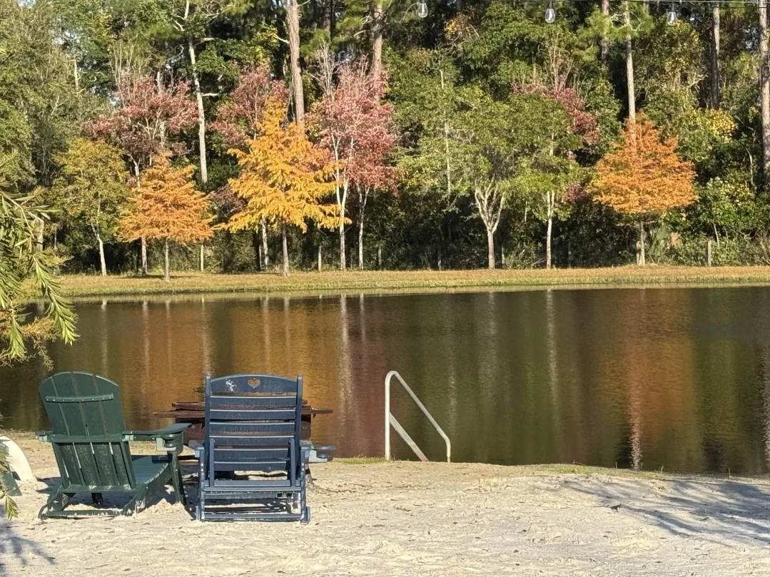Adirondack chairs and a glider seat on a sandy lakeside beach with autumn foliage reflecting on calm pond waters at Ruby Oaks Events wedding venue in Central Florida