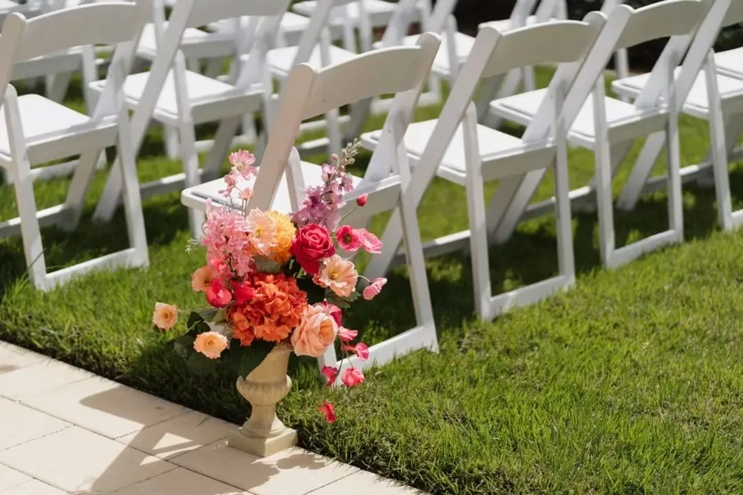 Colorful pink, coral, and orange floral arrangement in a white urn vase along the outdoor ceremony aisle with white folding chairs on green grass at Ruby Oaks Events garden wedding venue in Central Florida