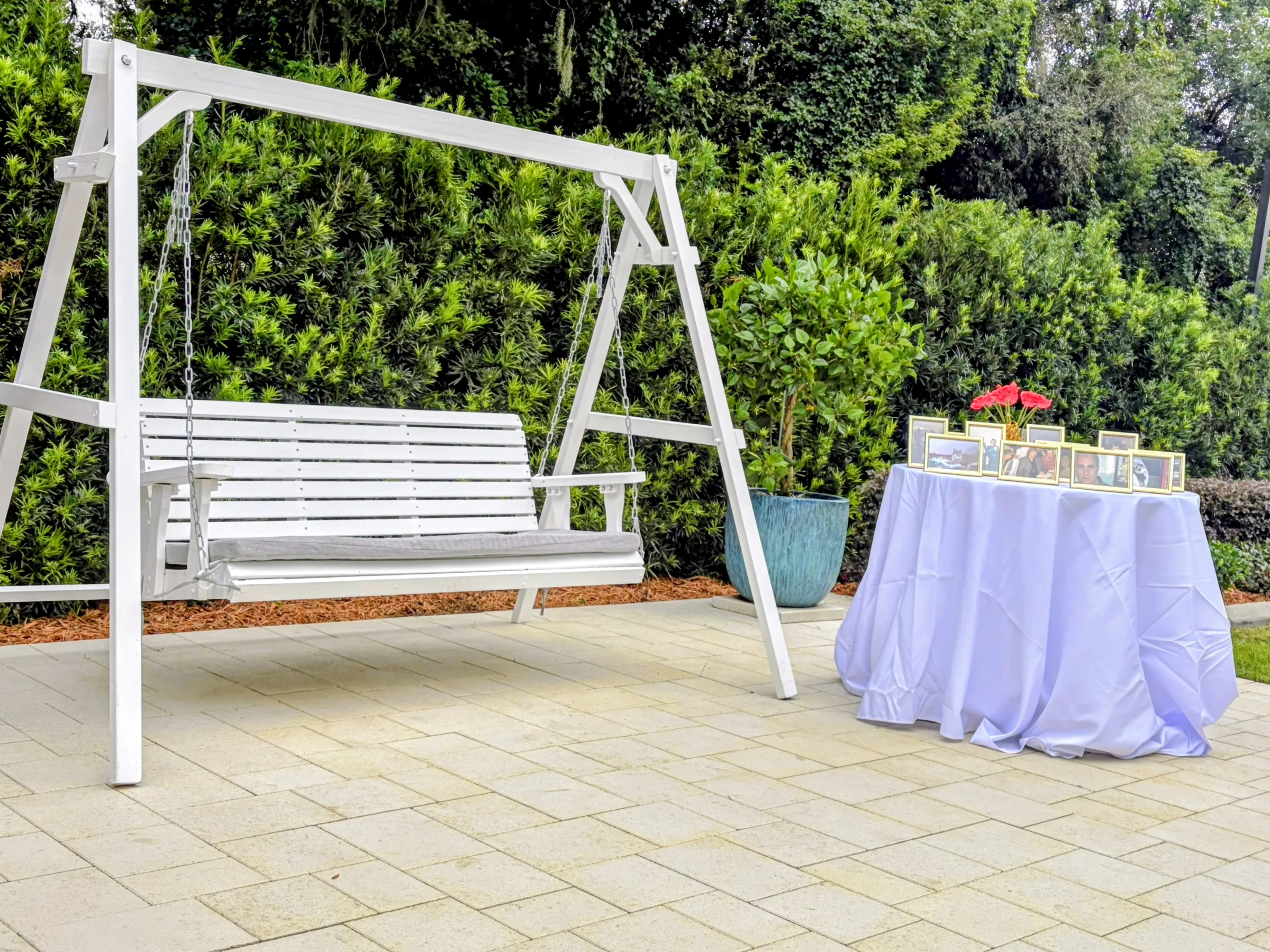 White wooden porch swing on a paver patio with a lavender-draped cocktail table displaying framed photos and red roses in the lush garden setting at Ruby Oaks Events wedding venue in Central Florida