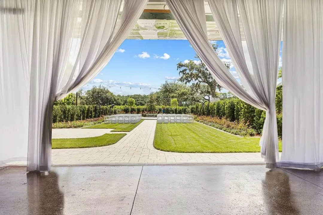 Outdoor garden wedding ceremony setup at Ruby Oaks Events in Central Florida, viewed through sheer white draped curtains from an indoor ballroom, featuring white folding chairs arranged along a paved aisle surrounded by lush greenery and mature oak trees under a bright blue sky