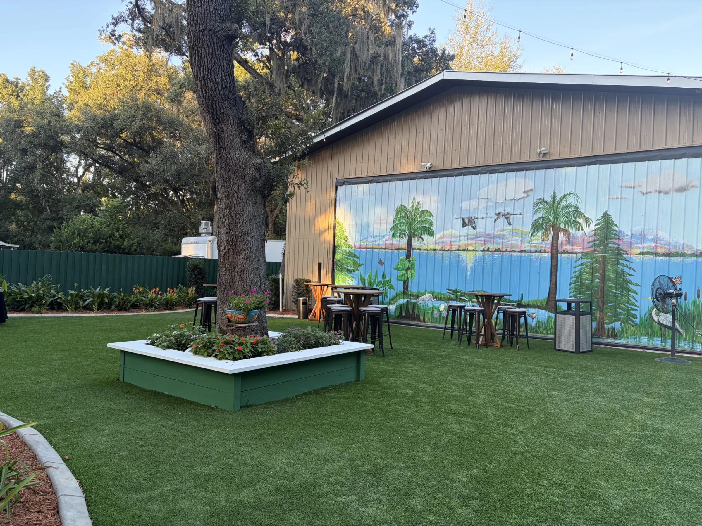 Outdoor garden patio at Ruby Oaks Events in Central Florida featuring artificial turf lawn, a raised garden planter surrounding a large oak tree, high-top bar tables and stools, and a colorful Florida nature mural painted on the exterior wall of the venue building