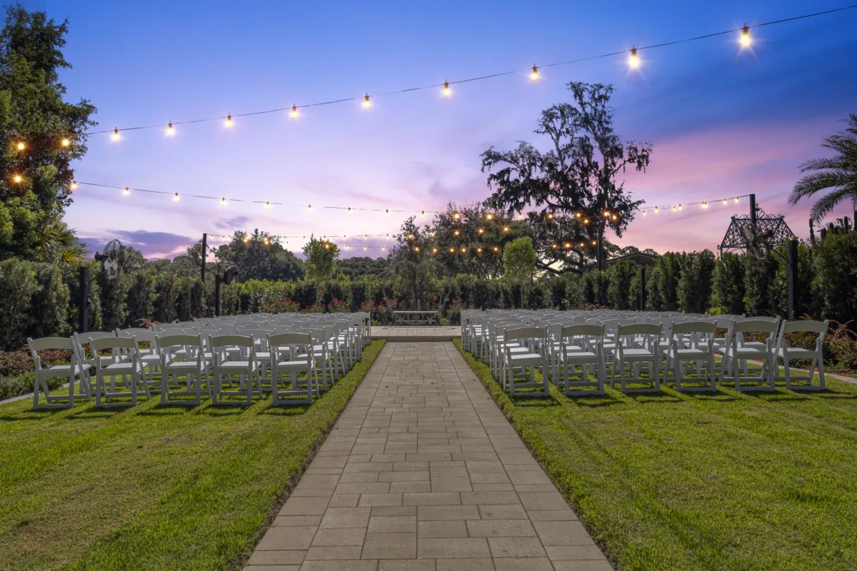 Outdoor garden wedding ceremony setup at Ruby Oaks Events in Central Florida featuring rows of white folding chairs along a paver aisle under string lights at twilight with a purple and pink sunset sky