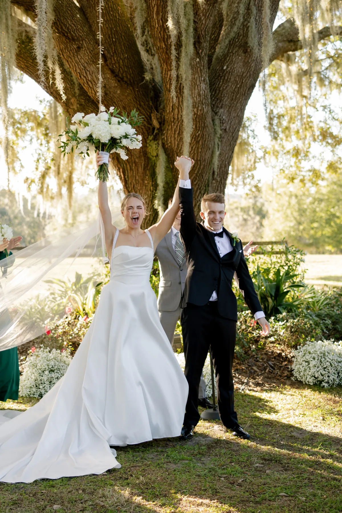 Bride and groom celebrate with raised hands and white bouquet after outdoor wedding ceremony under a large moss-draped oak tree at La Hacienda on Snow Hill in Tampa Florida