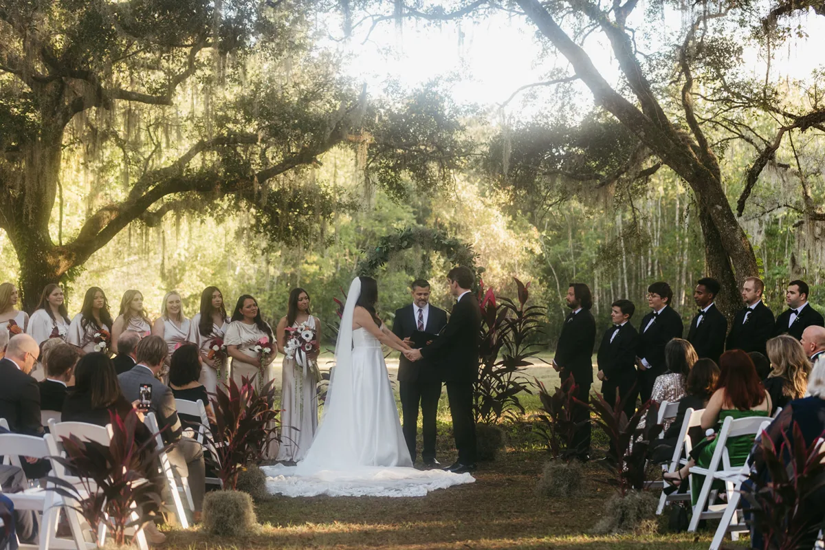 orange star farms 02da772a - Orangestar Farms Couple in wedding attire standing under tree branches draped in spanish moss with friends and family watching from an audience of white chairs.
