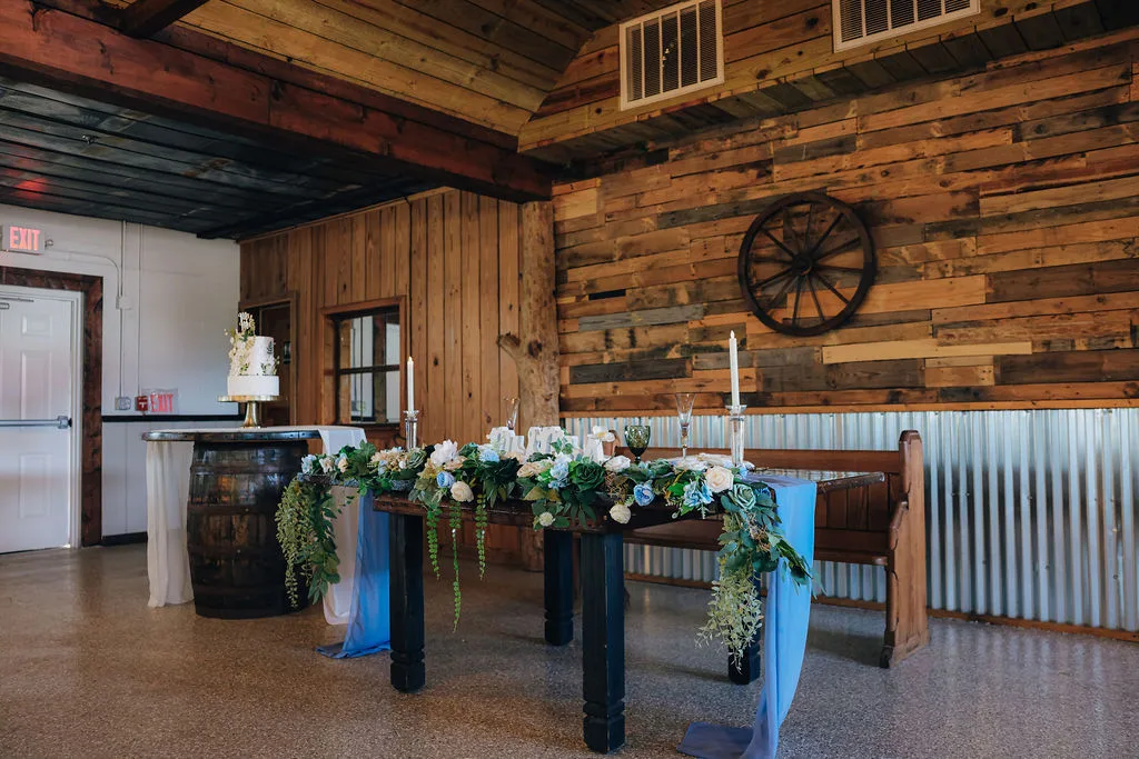 Large farmhouse sweetheart table at The Ranch at St. Cloud with floral garland, candles, and wood backdrop inside reception barn.