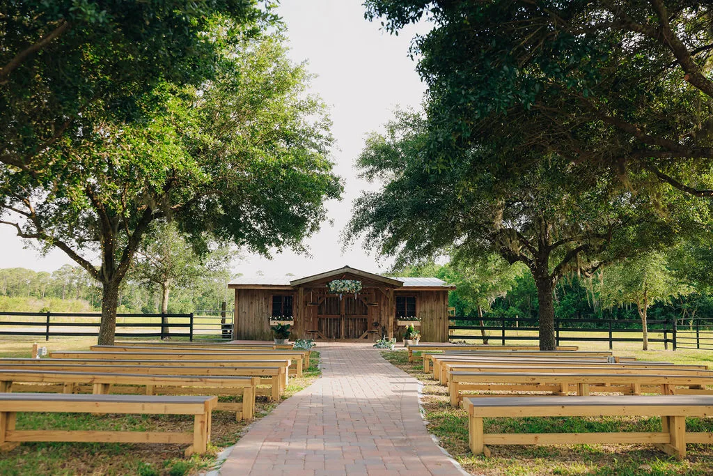 Outdoor wedding ceremony at The Ranch at St. Cloud Rustic Shack with wooden benches, brick aisle, and oak trees surrounding a barn-style backdrop.
