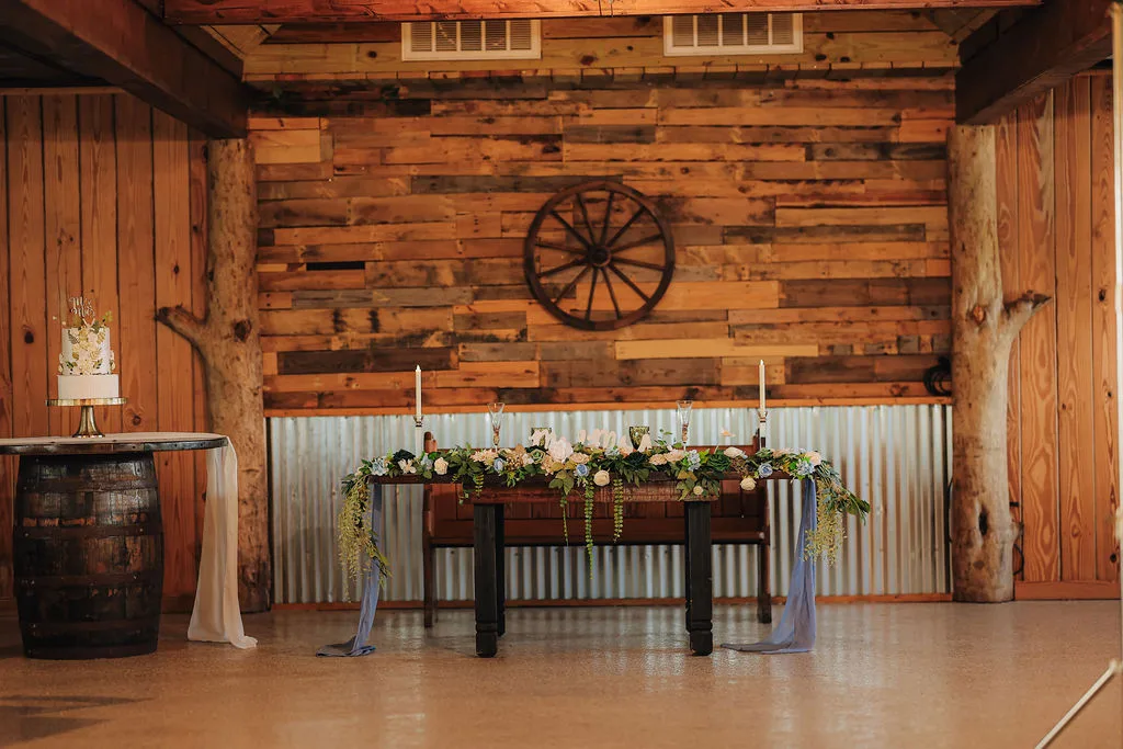 Decorated sweetheart table at The Ranch at St. Cloud with large farmhouse table, floral garland, candles, and rustic wood backdrop.