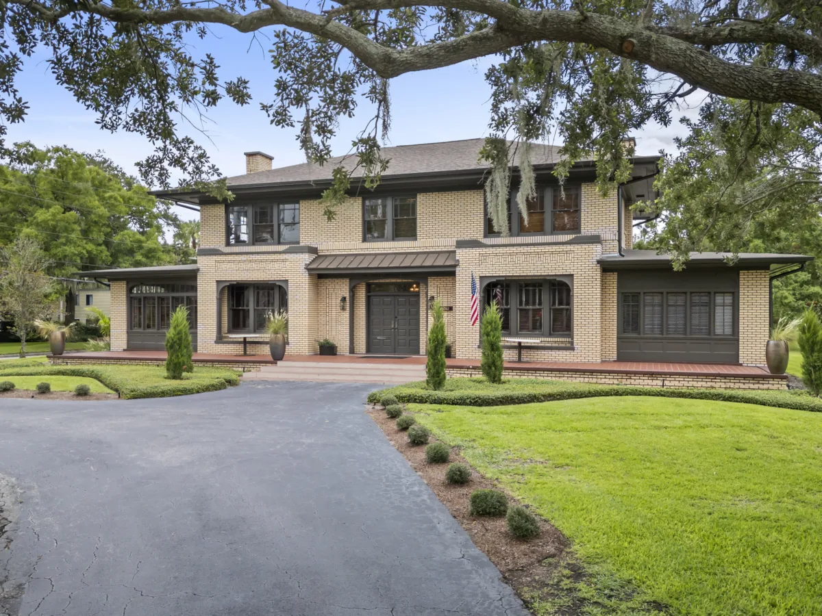 Large two-story historic brick building in downtown Orlando with a symmetrical façade, dark trim, and central front entrance, viewed from a wide driveway with ample on-site parking, manicured landscaping, hedges, and large oak tree branches overhead