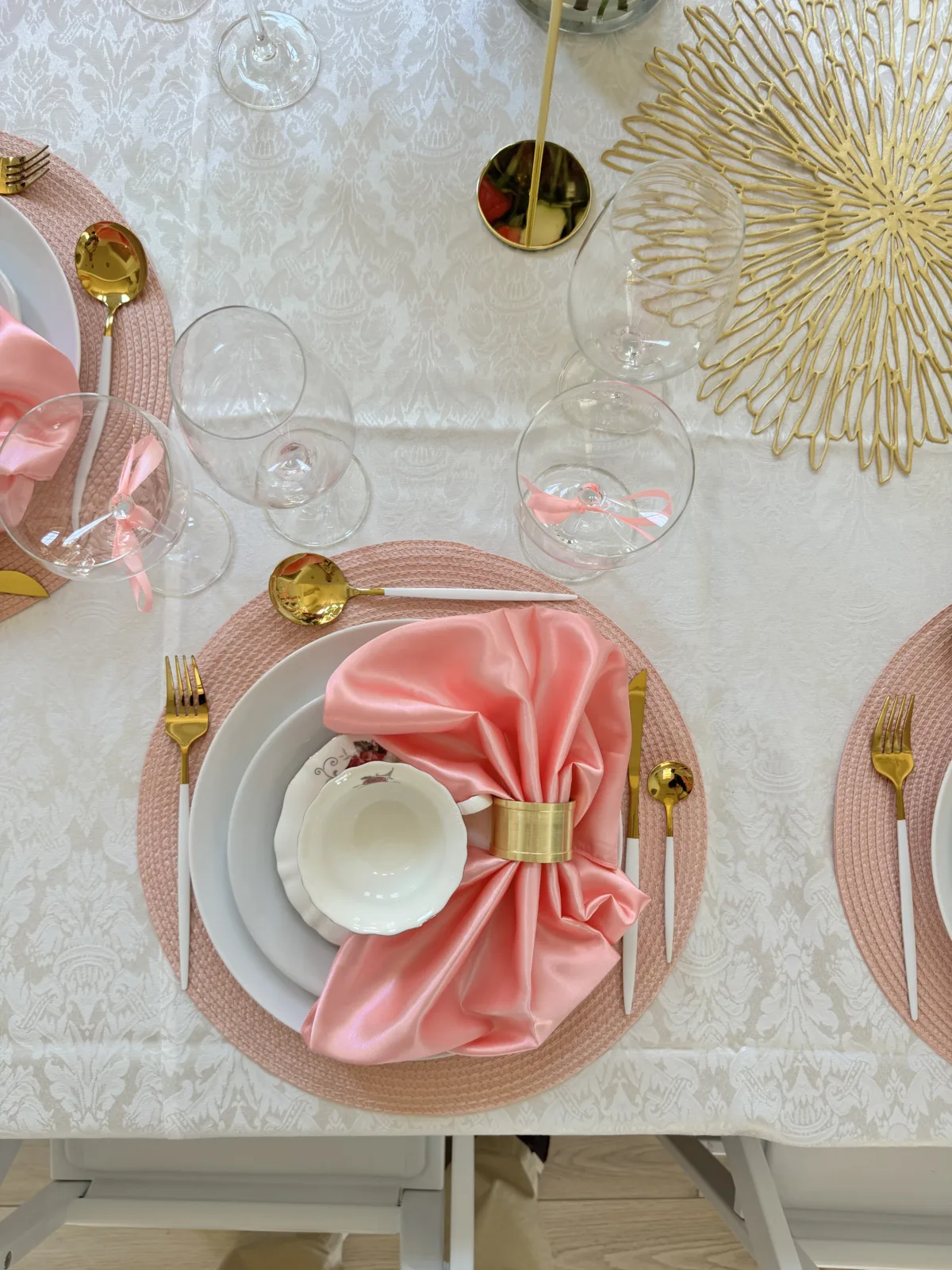 Elegant wedding table setting with blush pink woven placemats, gold cutlery, satin pink napkins with gold napkin rings, white china teacups, and crystal wine glasses on a white damask tablecloth at an Orlando Central Florida wedding