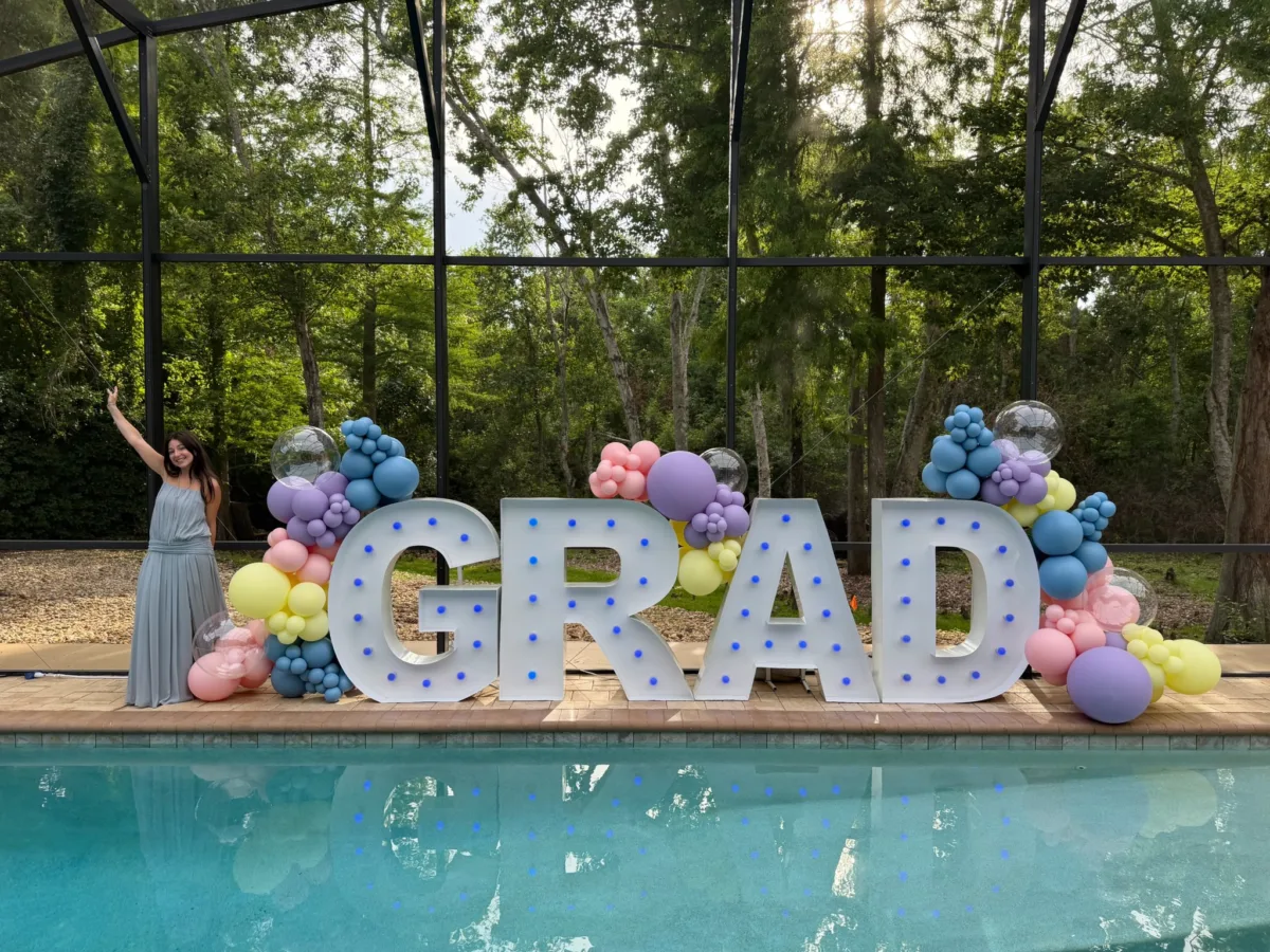 Illuminated GRAD marquee letters decorated with pastel balloon garlands beside a screened pool in Central Florida, with a graduate in a gray dress celebrating at a graduation party