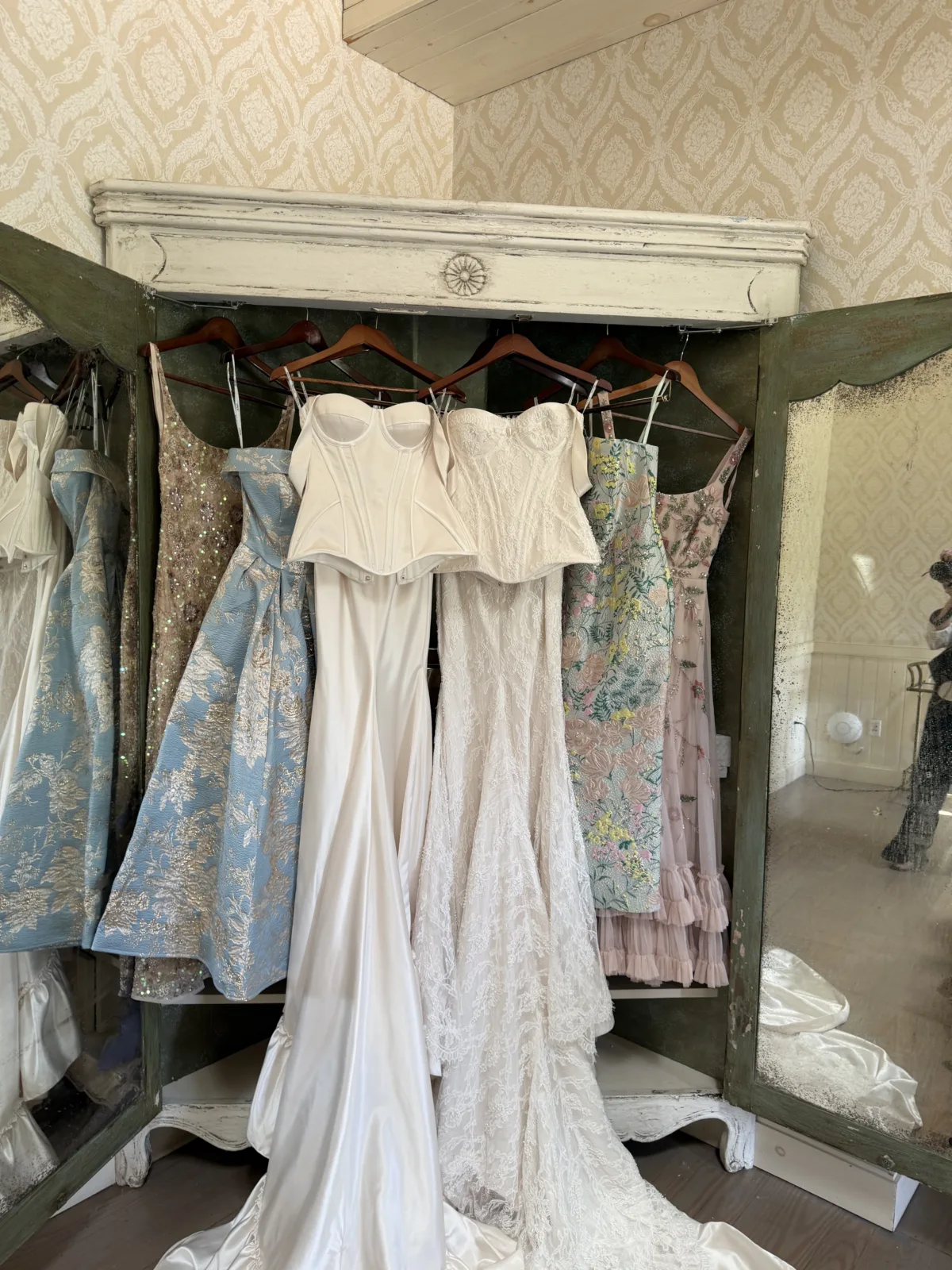 Wedding gown boutique display featuring white corset bridal gowns, a blue floral brocade dress, and colorful bridesmaid dresses hanging in a vintage distressed armoire wardrobe in Orlando Central Florida