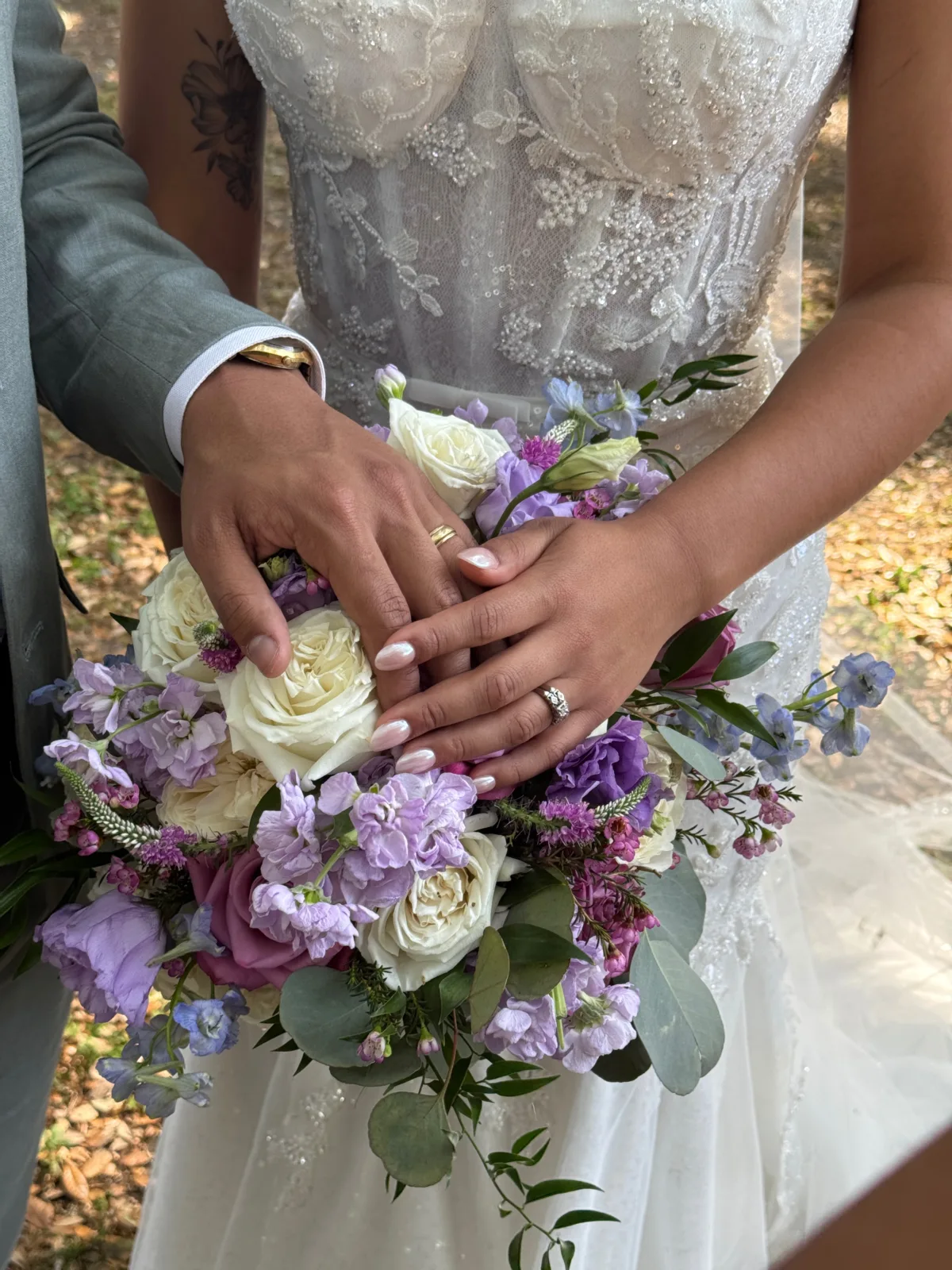 Bride and groom hands resting on a purple and white wedding bouquet featuring garden roses, lisianthus, stock flowers, blue delphinium, and eucalyptus, with wedding rings visible, at an Orlando Central Florida wedding