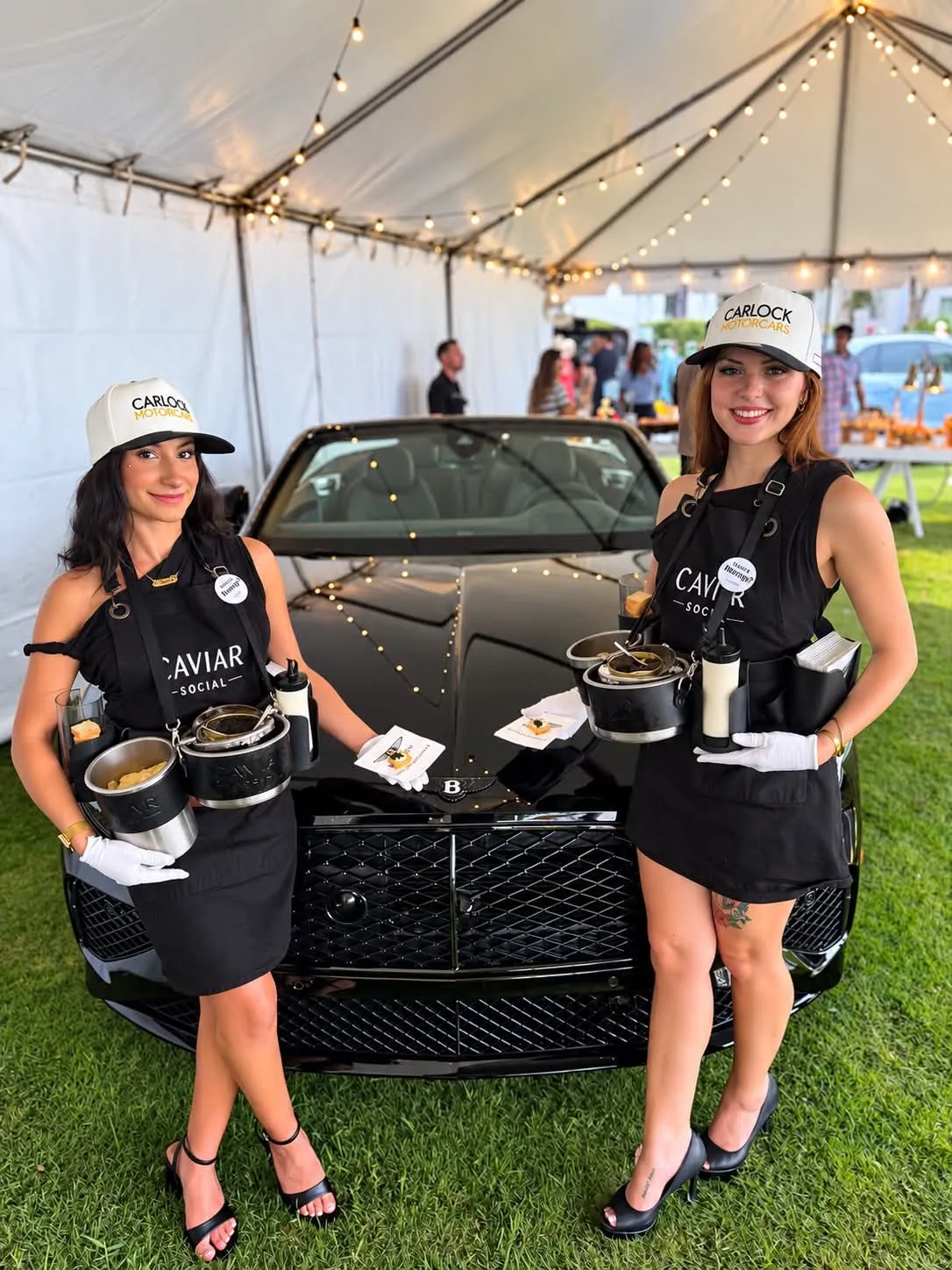 Caviar Social catering staff in branded black aprons and Carlock Motorcars caps serving caviar at an upscale outdoor event under a tent with string lights beside a black Bentley in Orlando Florida