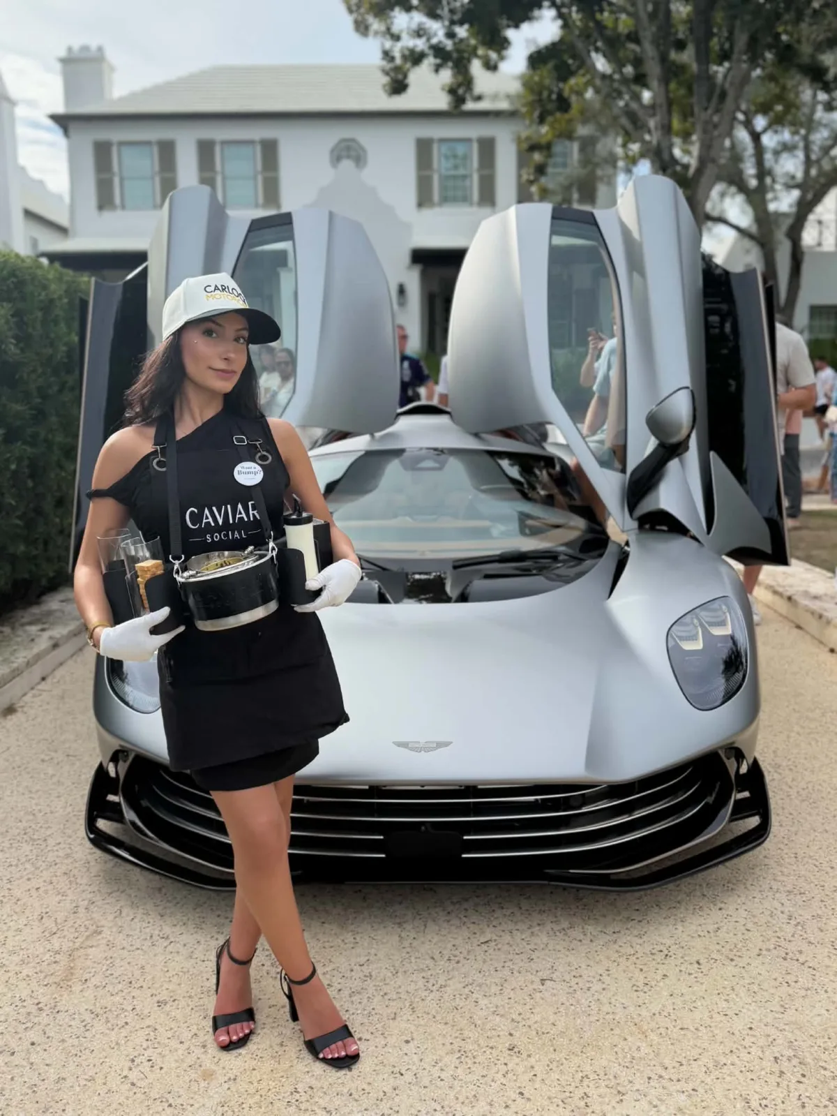 Caviar Social brand ambassador in black uniform and white gloves holding a caviar service tray in front of a silver Aston Martin with scissor doors open at a luxury event in Central Florida