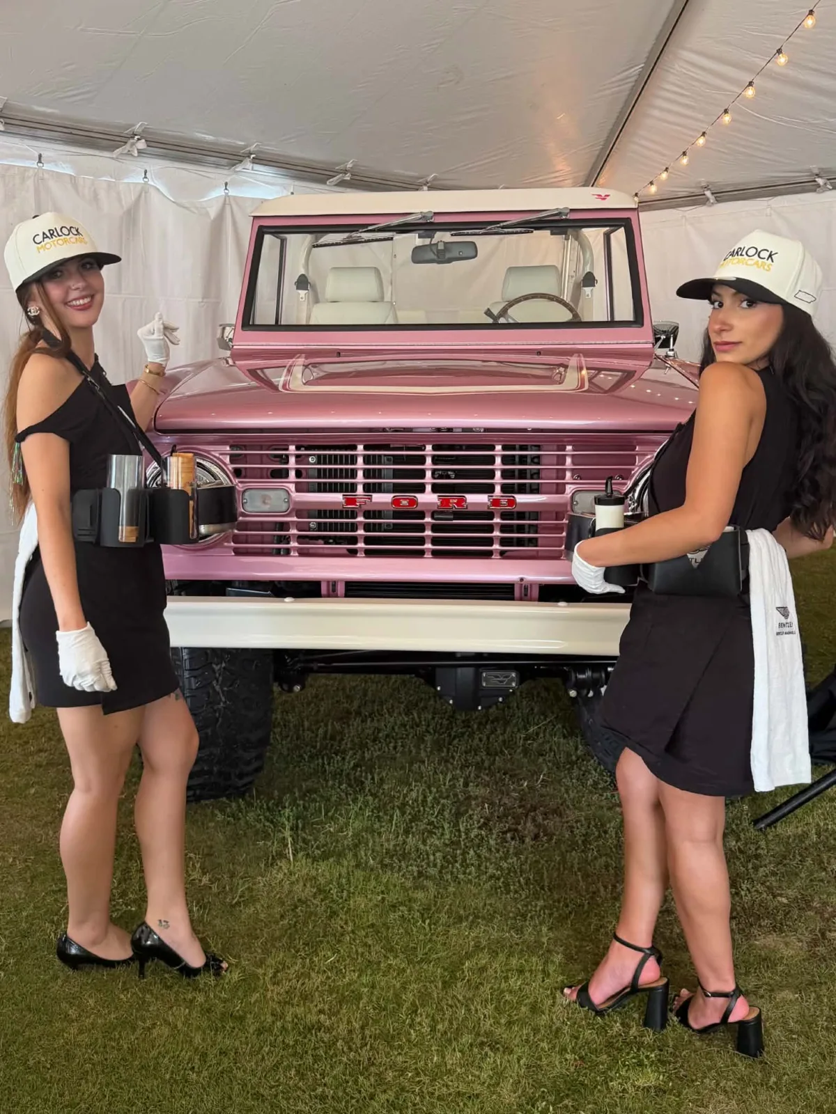Two brand ambassadors in black dresses and Carlock Motorcars hats pose in front of a custom pink vintage Ford Bronco at a Caviar Social luxury event experience in Orlando Florida