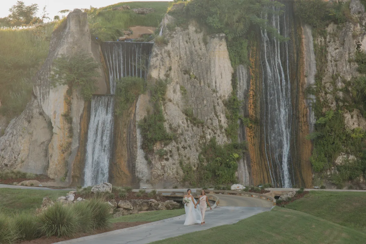 Two women in formal wedding attire walk hand in hand along a stone pathway in front of a dramatic limestone cliff waterfall at The Club at Iron Lake wedding venue in Ocala Florida