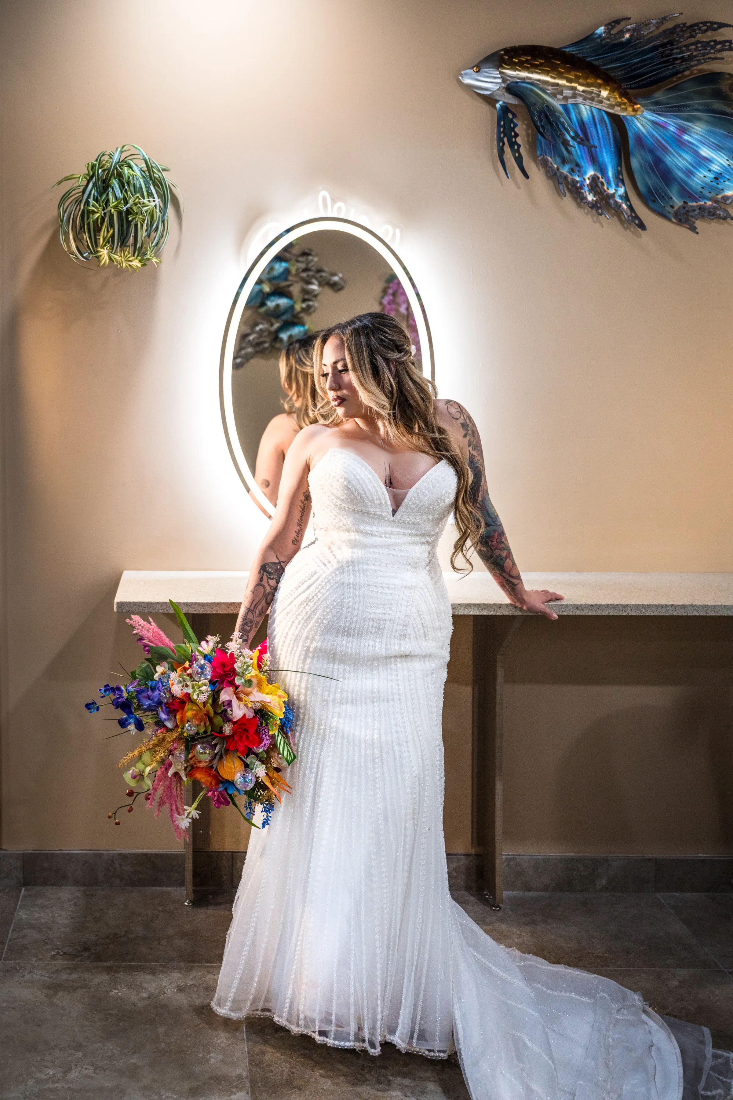 Tattooed bride in a strapless beaded white wedding gown holding a vibrant tropical bouquet of colorful mixed flowers while posing in front of an illuminated oval mirror at Daytona Aquarium in Daytona Beach Florida during a styled shoot wedding inspiration session
