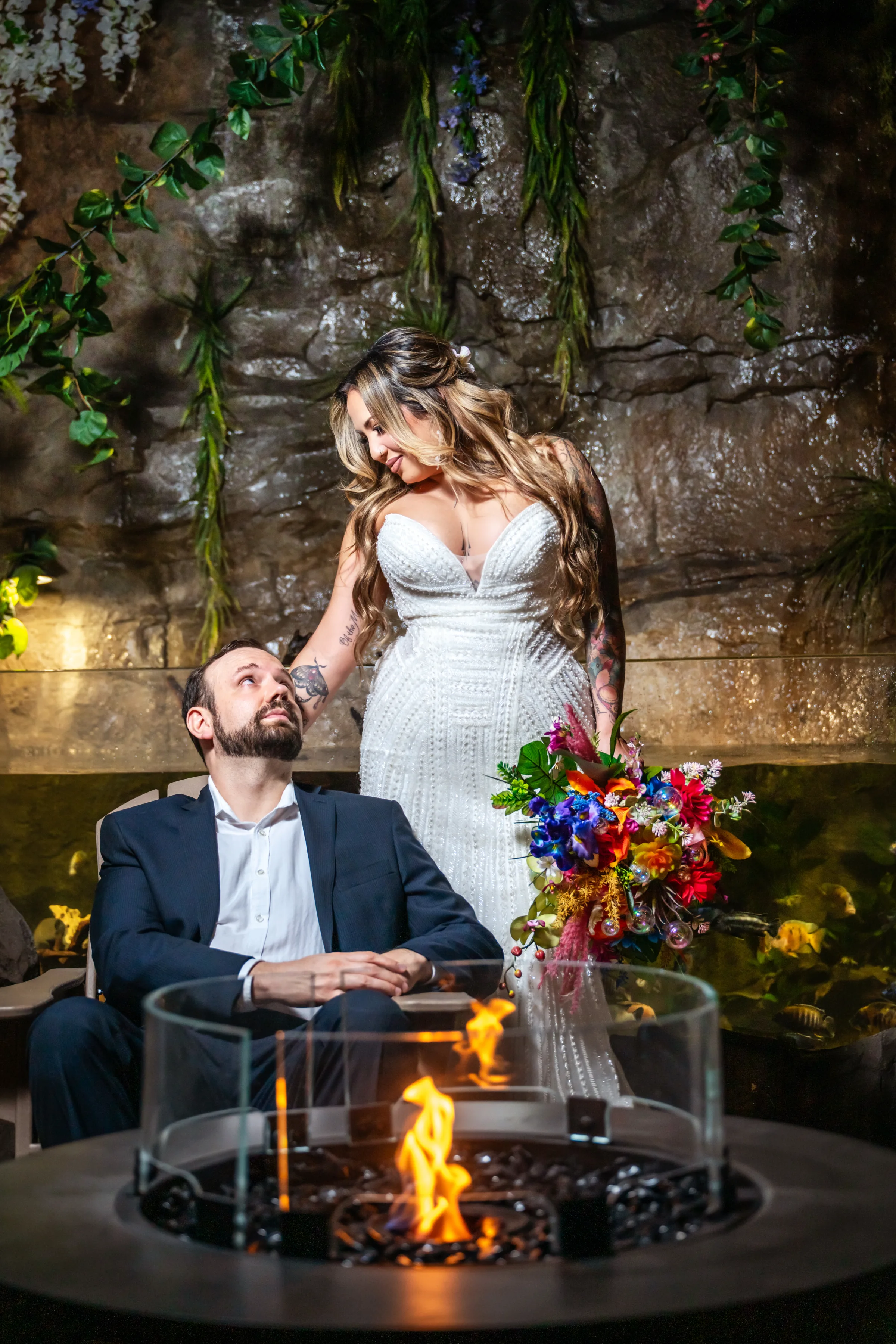 Bride in a beaded strapless gown holding a vibrant tropical bouquet stands beside a seated groom in a navy suit near a fire pit, with hanging greenery and a rocky waterfall wall backdrop at Daytona Aquarium in Daytona Beach Florida