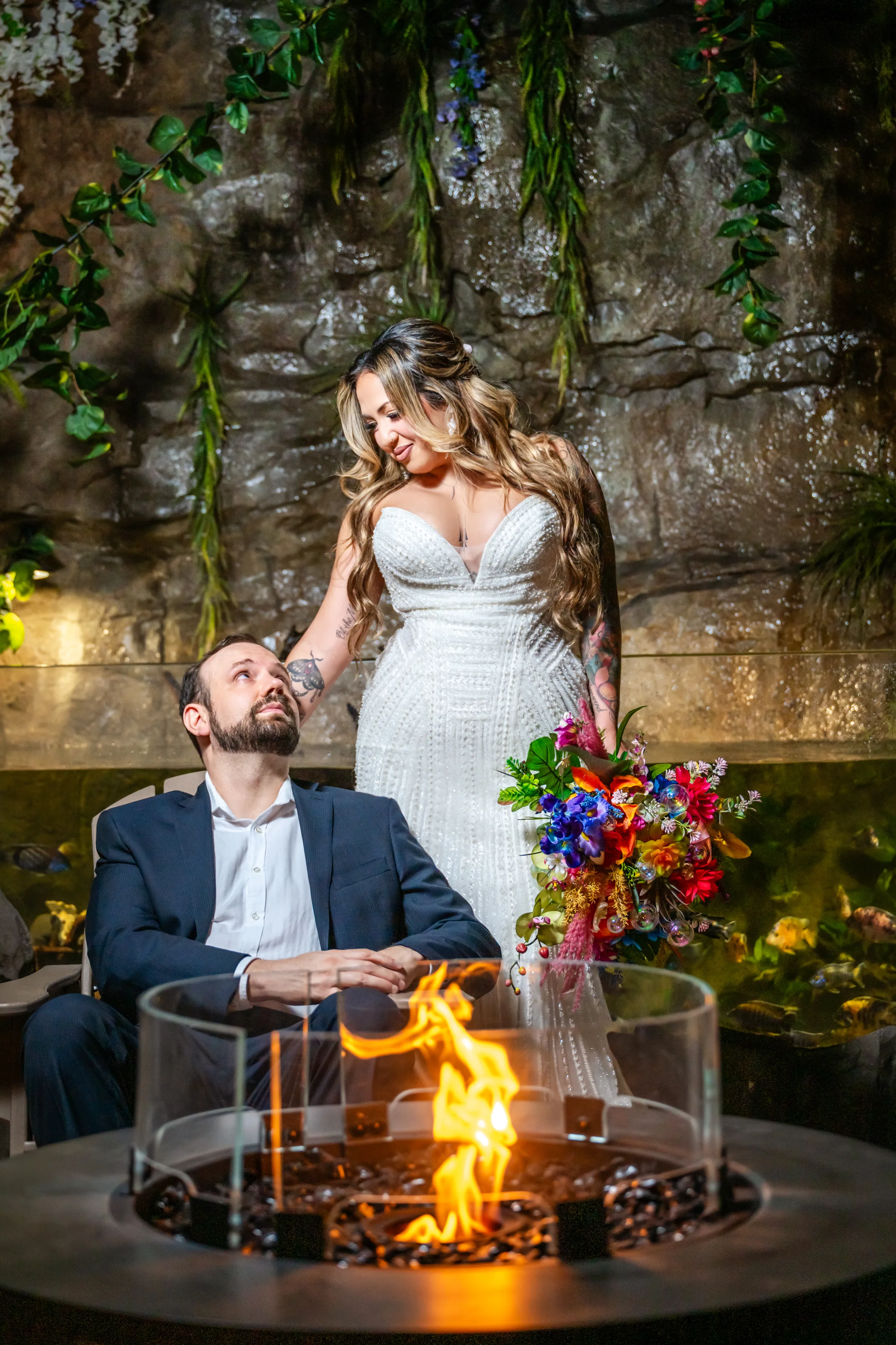 Bride in a beaded strapless gown holding a vibrant tropical bouquet stands beside a seated groom in a navy suit near a fire pit at Daytona Aquarium in Daytona Beach Florida during a styled shoot wedding inspiration session