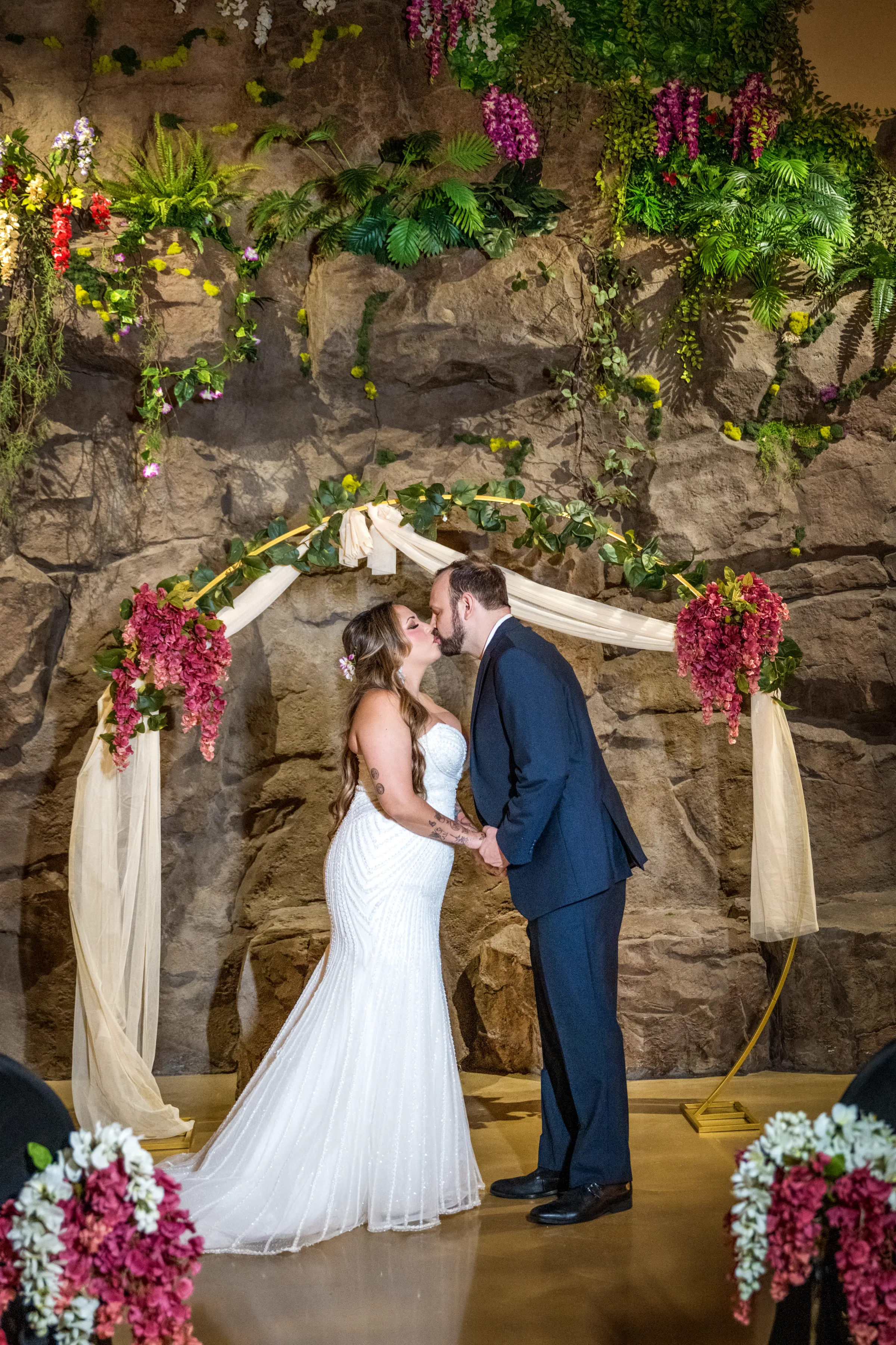 Bride and groom share their first kiss under a floral arch adorned with cascading pink flowers and ivory draping against a dramatic rock wall backdrop at Daytona Aquarium in Daytona Beach Florida during a tropical aquarium wedding ceremony
