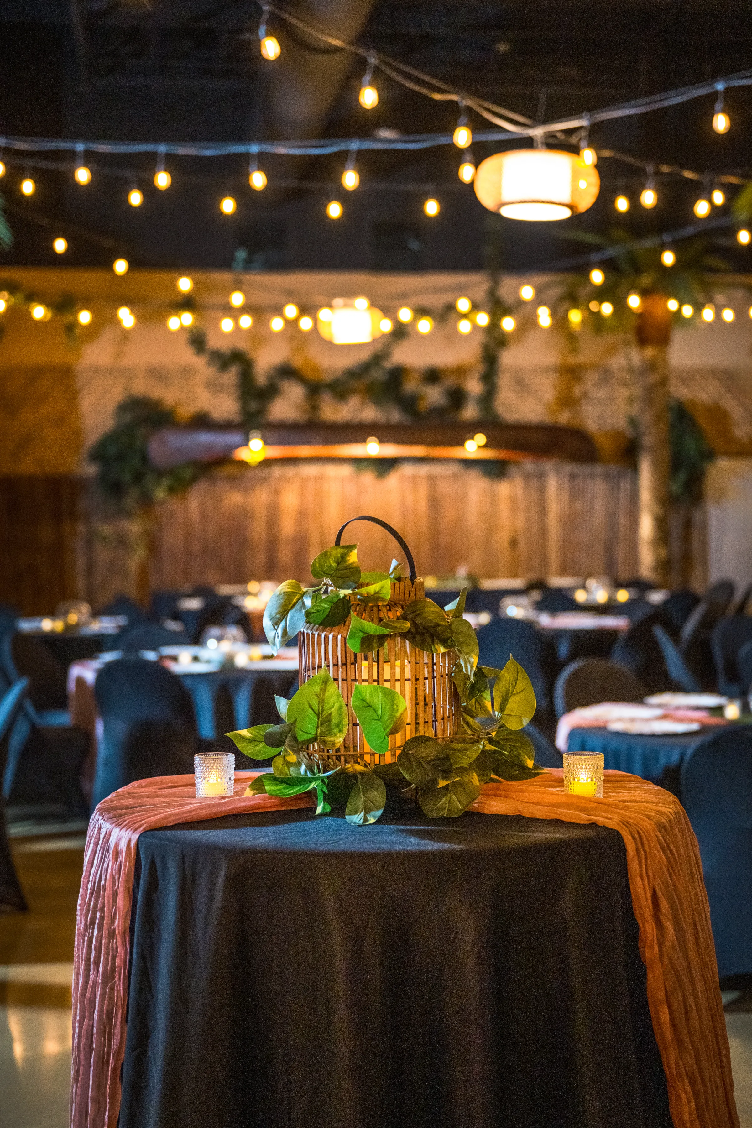 Tropical wedding reception tablescape at Daytona Aquarium featuring a bamboo lantern centerpiece wrapped in lush green tropical leaves on a navy linen with a terracotta runner, flanked by votive candles, under warm string lights in an intimate aquarium event space in Daytona Beach Florida