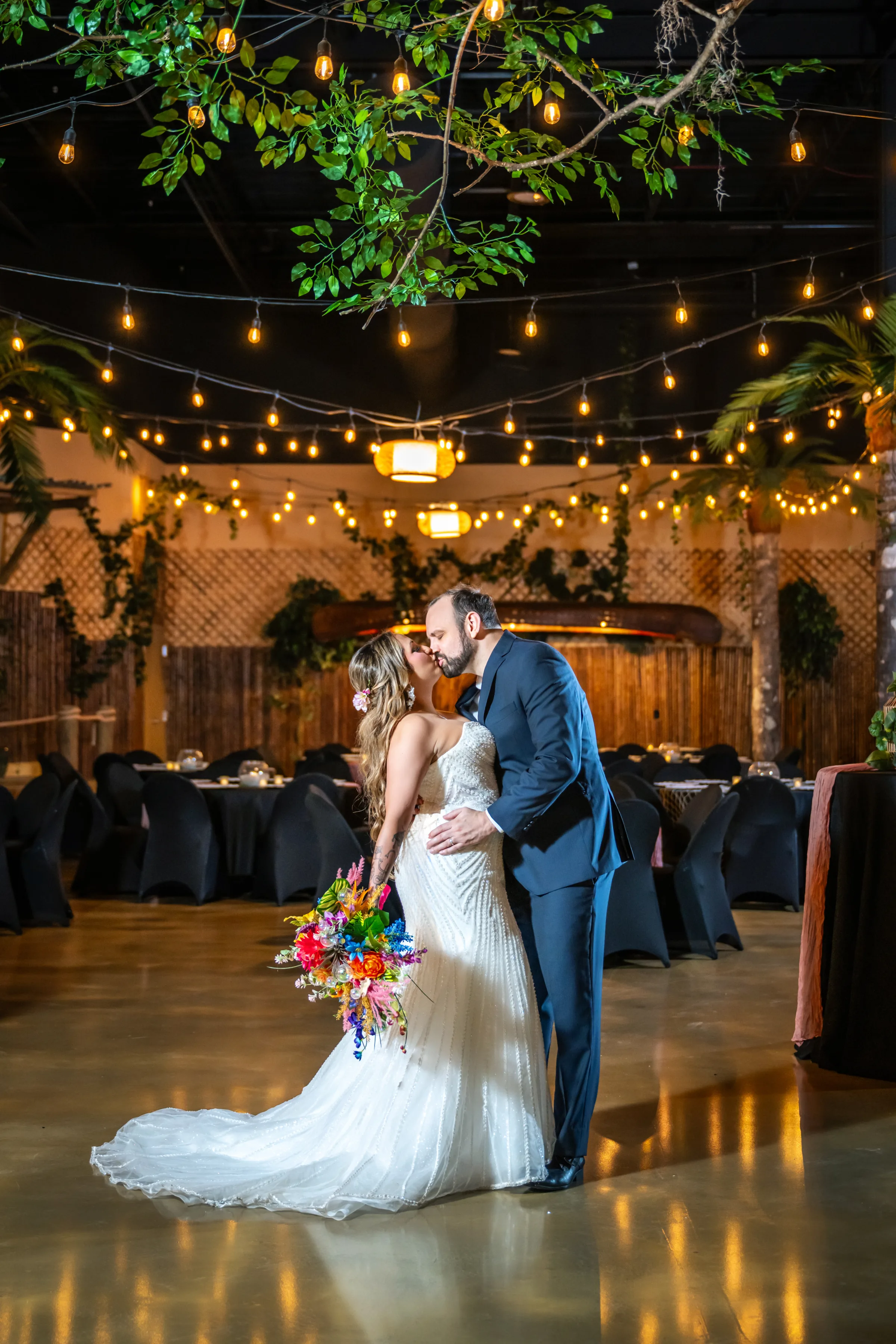 Bride and groom sharing a kiss on the dance floor at Daytona Aquarium during a tropical wedding styled shoot, surrounded by string lights, hanging greenery, and black draped reception tables, bride holding a vibrant colorful bridal bouquet