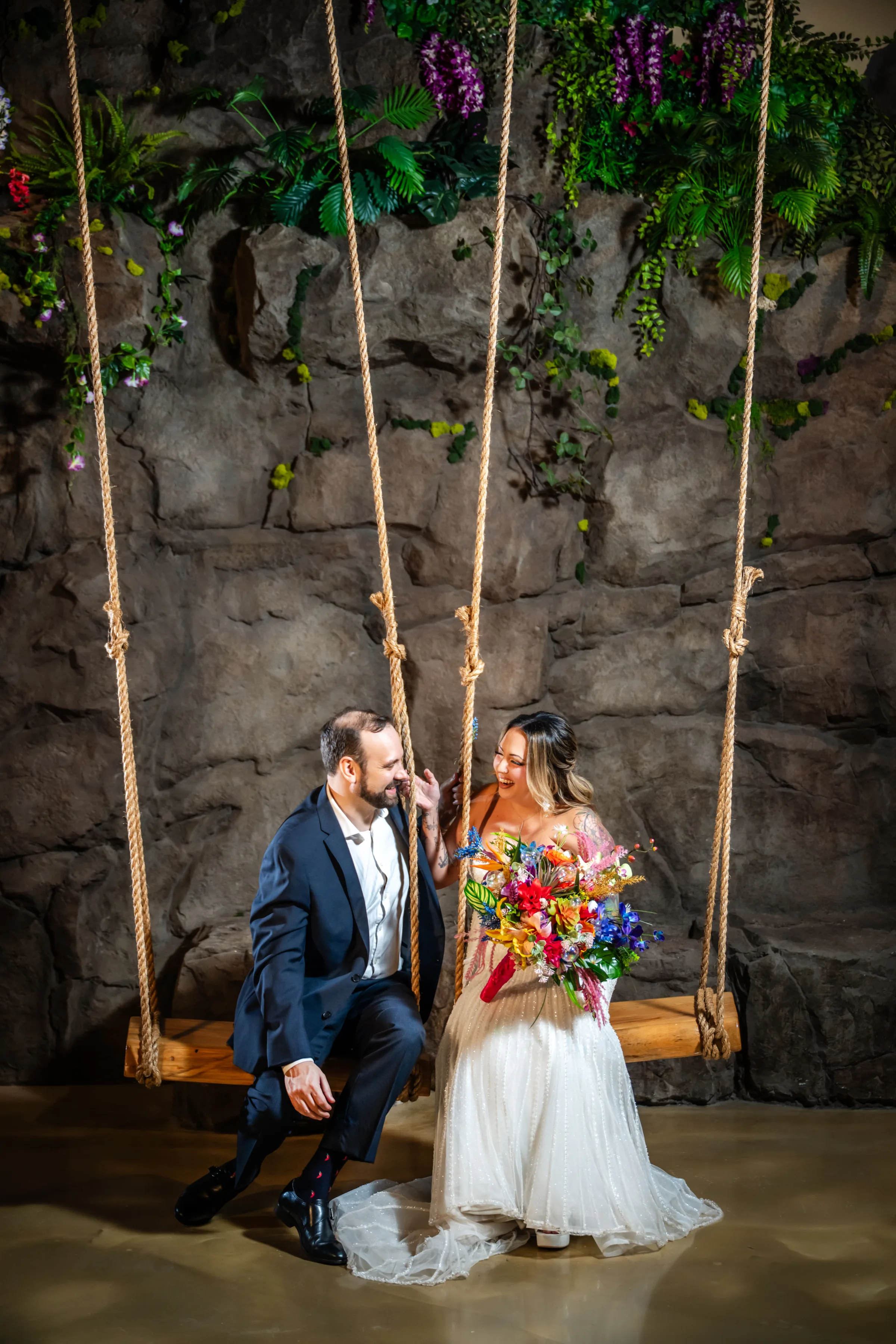 Bride and groom sitting together on a rope swing bench in front of a stone wall with lush tropical greenery and purple flowers at Daytona Aquarium, bride holding a vibrant multicolored bouquet in a sparkly floral wedding gown