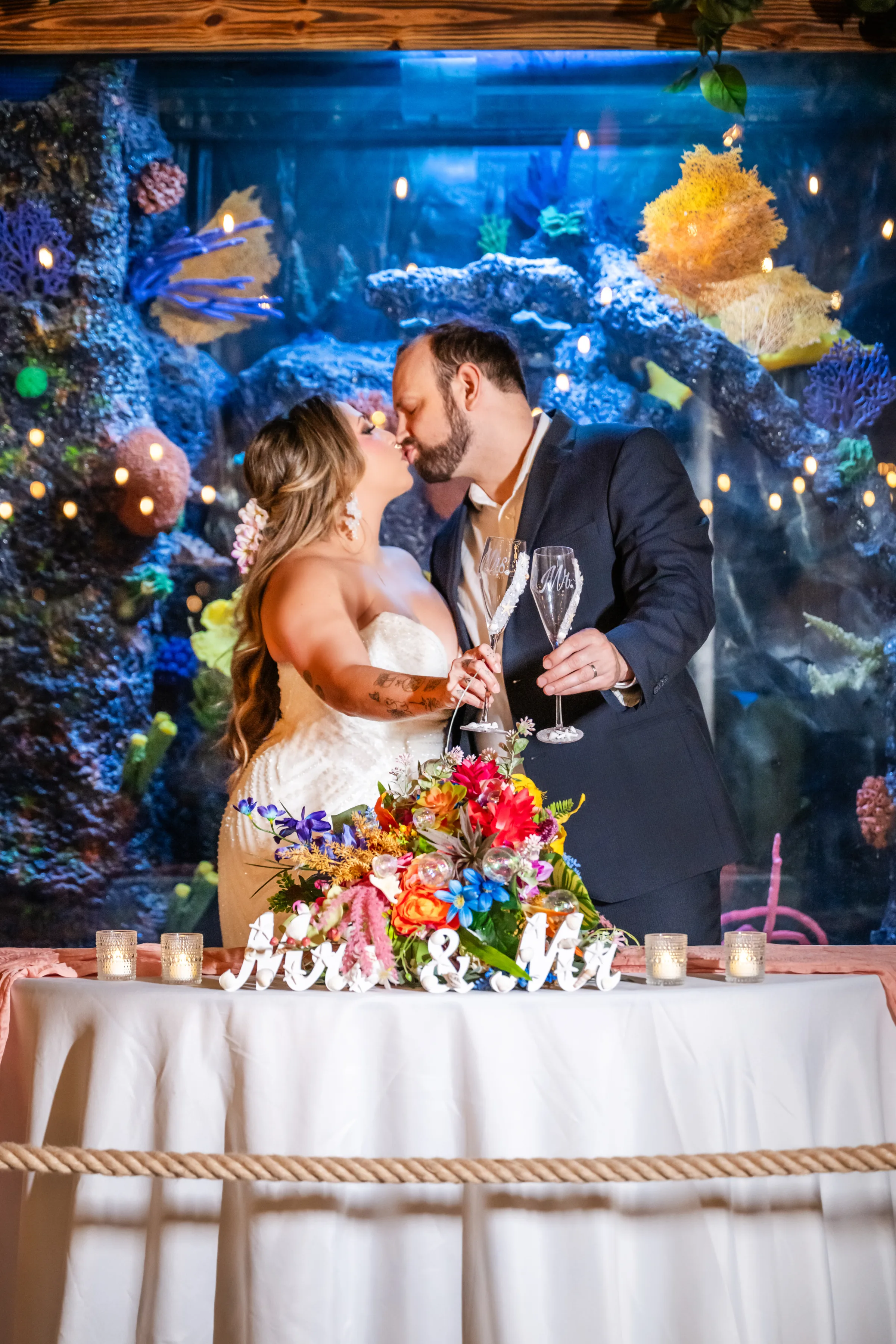 Bride and groom share a kiss at the sweetheart table in front of a vibrant coral reef aquarium display at Daytona Aquarium, holding champagne flutes engraved with Mr and Mrs, with a colorful tropical floral centerpiece and Mrs and Mr table sign at this unique alternative wedding venue in Daytona Beach Florida