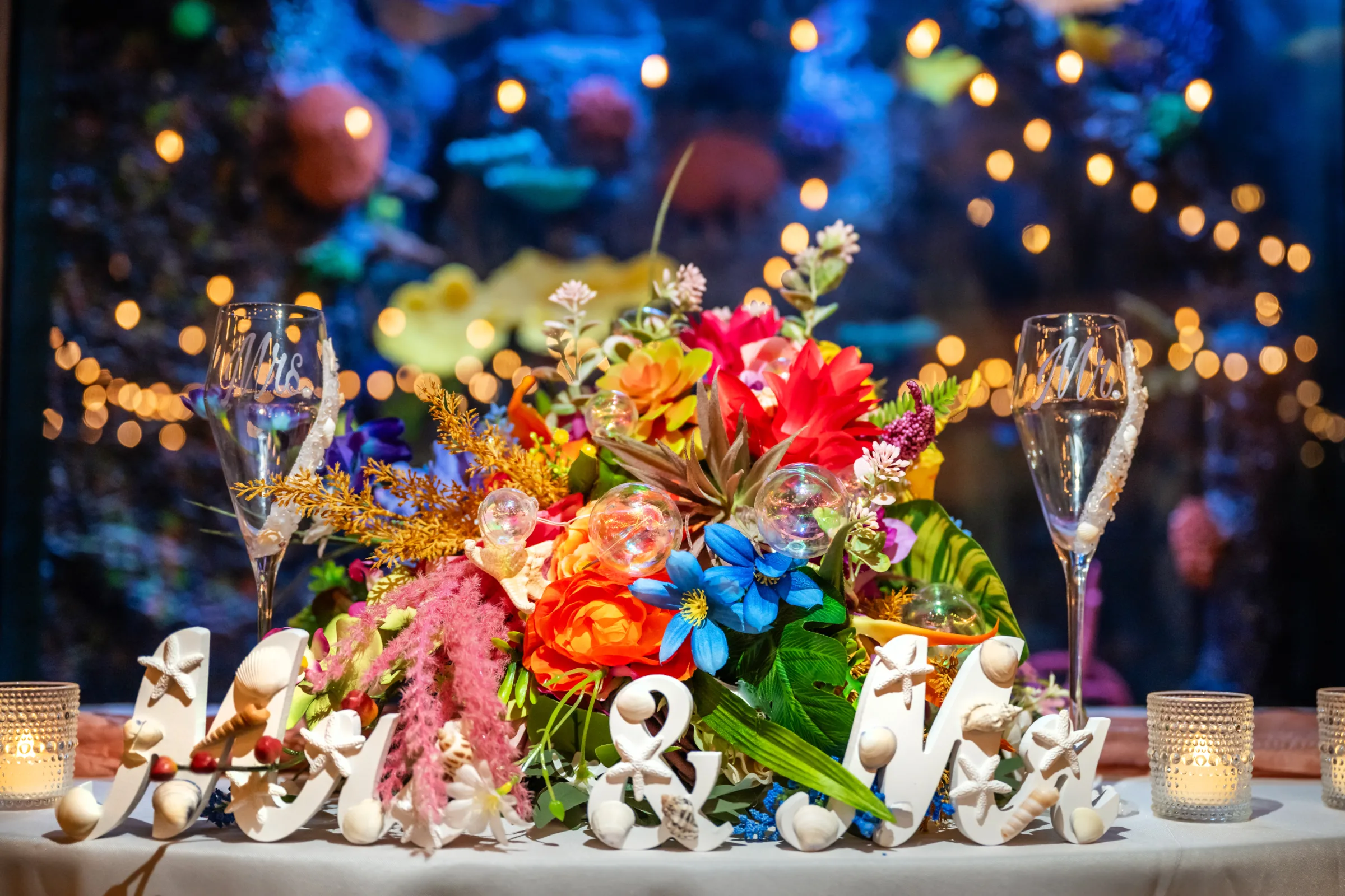Colorful tropical wedding sweetheart table centerpiece with vibrant rainbow florals, Mr. and Mrs. champagne flutes, seashell name signs, and candlelight at Daytona Aquarium in Daytona Beach Florida