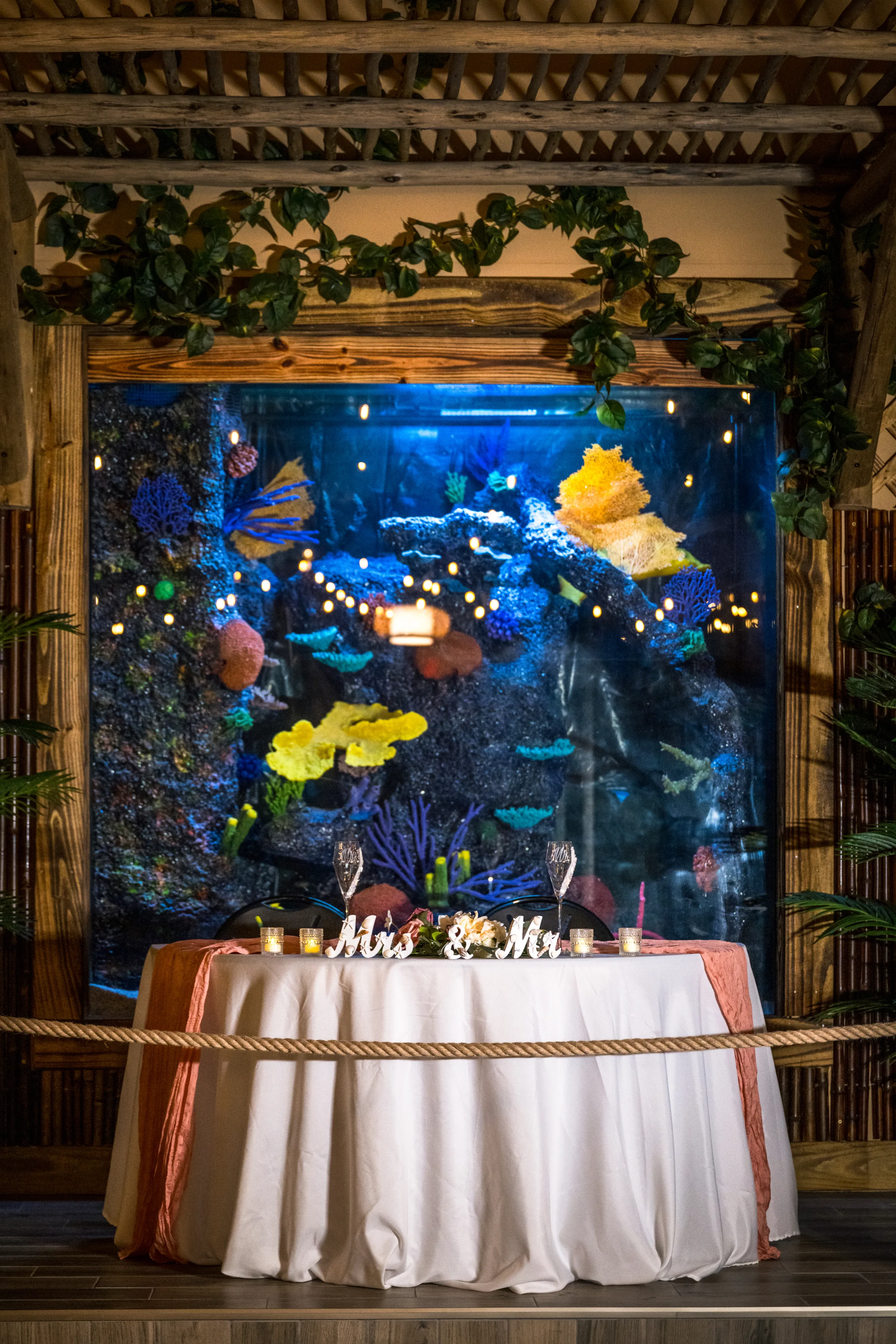 Mrs and Mr sweetheart table with white linen and coral runner set in front of a large illuminated coral reef aquarium tank at Daytona Aquarium, a unique tropical wedding venue in Daytona Beach Florida